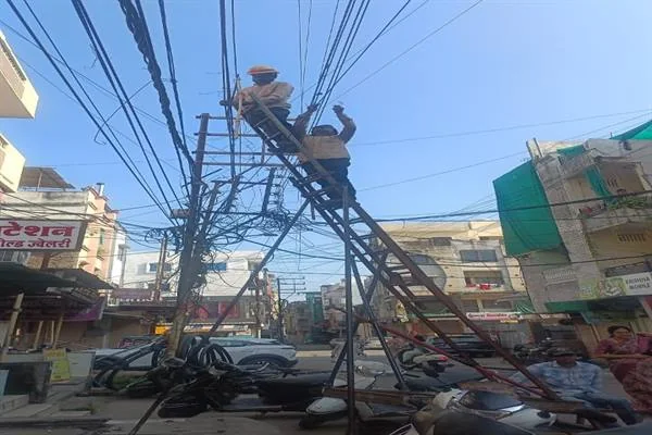 तारों से घिरे बिजली के खंभे पर सीढ़ी चढ़कर काम करते दो बिजलीकर्मी