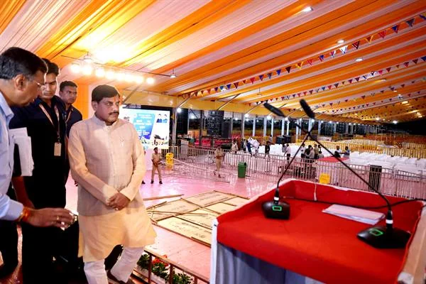 मुख्यमंत्री डॉ. मोहन यादव सफेद कुर्ते और आसमानी जैकेट में पंडाल में बने मंच के पास खड़े होकर व्यवस्थाएं देख रहे हैं; आगे लाल कपड़े से ढकी मेज पर माइक लगे हैं और पीछे कुर्सियों से सजा सभा स्थल दिख रहा है।